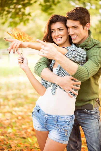 Молодая пара обнимается в парке протянутыми пальцами руки, указывающими на что-то, а девушка несет в руке веточку осенних листьев.
