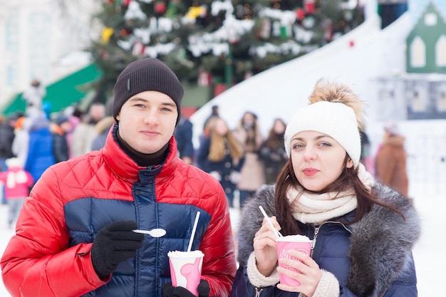 寒い雪の冬の日にクリスマスカーニバルを楽しんでいる若いカップルは、人々の群衆と背景に飾られた松の木とテイクアウトコーヒーを飲みながら屋外に立っています