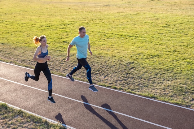 Young couple of fit sportsmen boy and girl running while doing exercise on red tracks of public stadium outdoors.