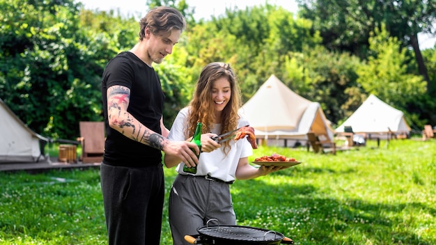 Young couple frying meat on the grill and drinking beer. Greenery around. Glamping