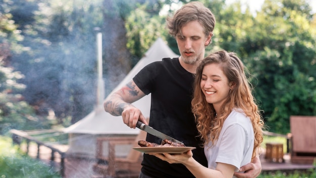 Young couple frying meat on the grill and putting it on the plate. Greenery around. Glamping