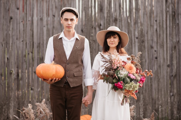 A young couple gently holds hands on each other. A man stands with a pumpkin in his hands, and a girl with a bouquet. Autumn environment