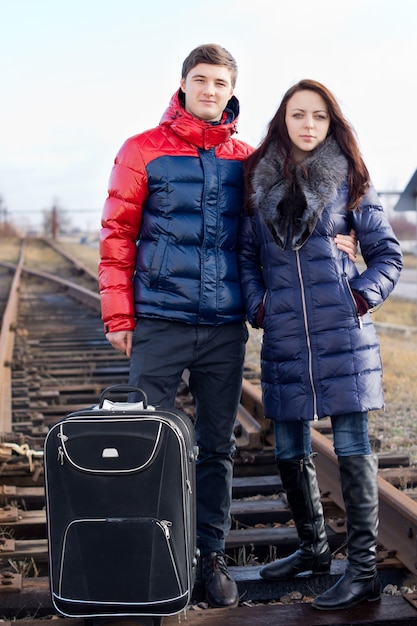 Young couple going off on vacation standing arm in arm with their suitcase in the centre of the train track on the wooden sleepers waiting for the train