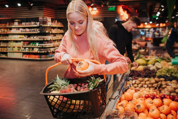 Young couple in grocery store. Nice woman put persimmon into grocery basket and look down.