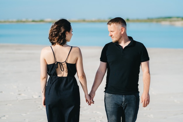 Young couple a guy with a girl in black clothes are walking on the white sand at the edge of blue water