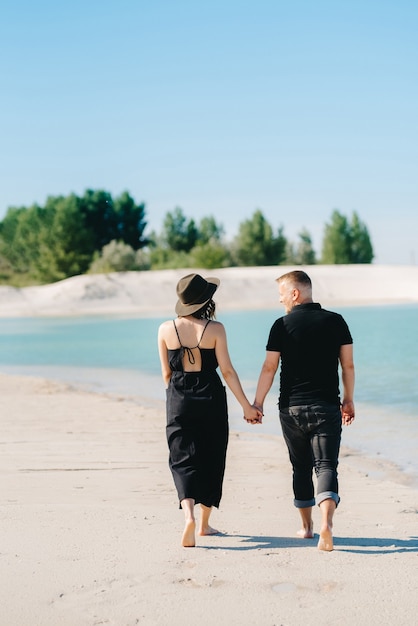 若いカップルは黒い服を着た女の子と男が青い水の端にある白い砂の上を歩いています