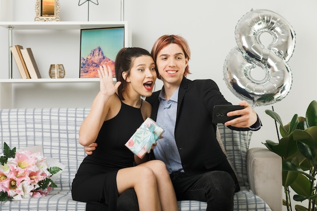 Young couple on happy women day holding present take a selfie sitting on sofa in living room