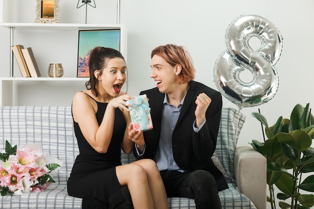 Young couple on happy women day pleased guy gives present to\
surprised girl sitting on sofa in living room