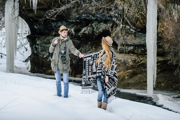 Young couple in hats is walking and smiling next to frozen river against background of rocks during winter walk.