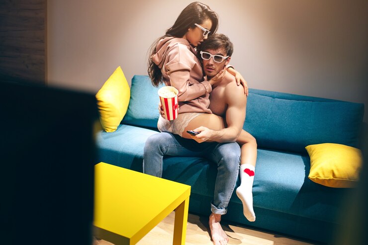 Photo young couple have intimacy in kitchen in night. hot seductive and sensual woman sit on guy's legs in sex position. shirtless man hold hands on her buttocks and watching tv. movie. concentrated