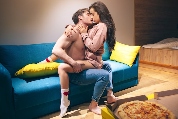 Young couple have intimacy in kitchen in night. Passionate shirtless man hold woman on his legs and kissing her. Seductive sensual model in sex position enjoying. Pizza on table.