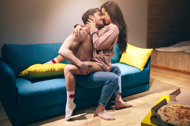 Young couple have intimacy in kitchen in night. Passionate shirtless man sit on sofa and kiss sensual woman.