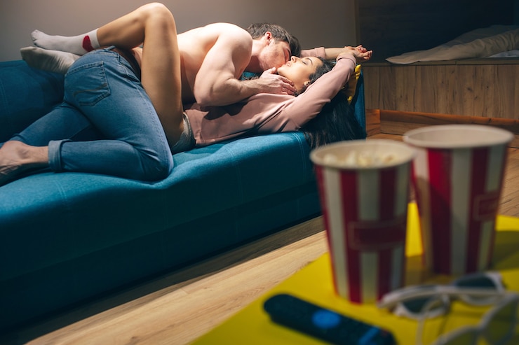 Photo young couple have intimacy in kitchen in night. shirtless man lying on seductive woman in sex position. model stretch hands and enjoy. popcorn buckets, cinema glasses and remote control on table.