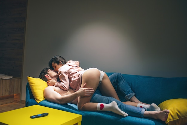 Young couple have intimacy in kitchen in night. Standing in sex position on sofa. Hot seductive woman on top. Shirtless man lying on sofa and hold hand on her hips. Sensual moment