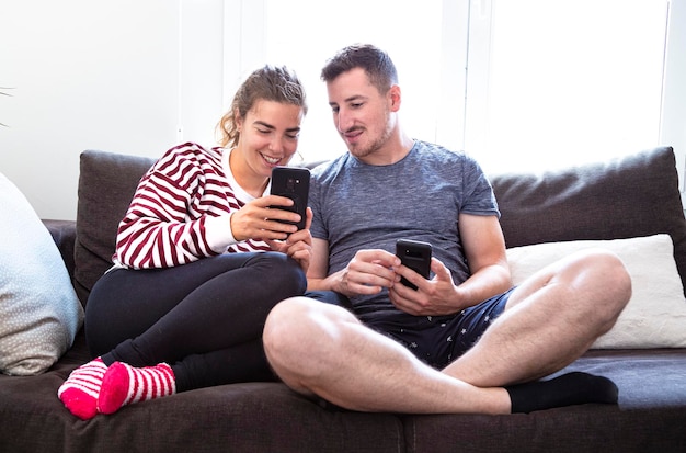 Young couple having fun and laughing interacting with their cell phones sitting on the couch at home