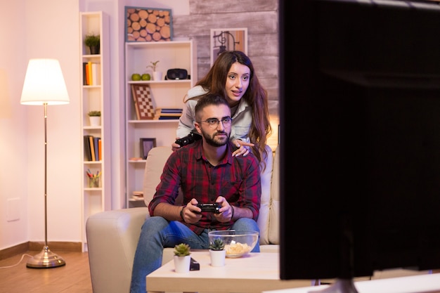 Young couple having fun while playing video games on televison sitting on couch at night.