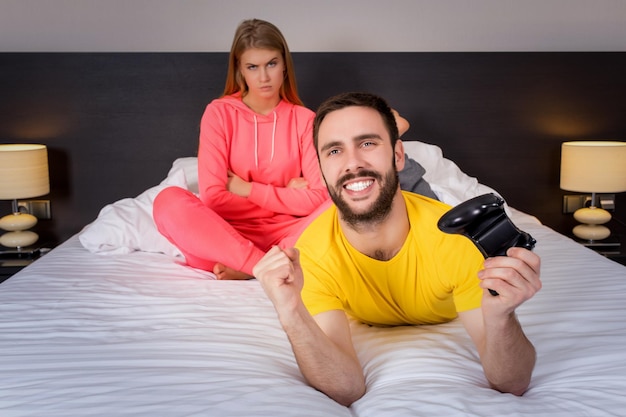 ベッドでビデオゲームをしている若いカップル。ビデオゲームをプレイしている男性、背景に動揺している女性。