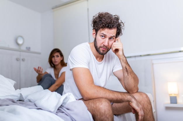 Photo young couple having a problem. handsome man is leaning on his hand while sitting sadly on bed, woman is sitting in the background. head full of thoughts when she is around