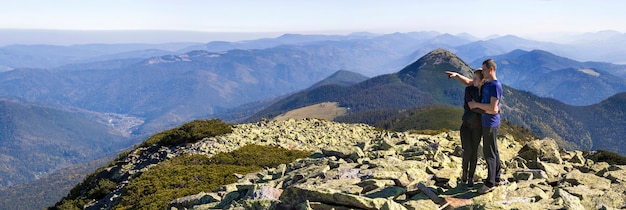 Young couple hike in Carpathian mountains. Man and woman standing on mountain top looking at beautiful landscape below. Wide panorama of high rocky hills in warm summer weather. Sports and activity.