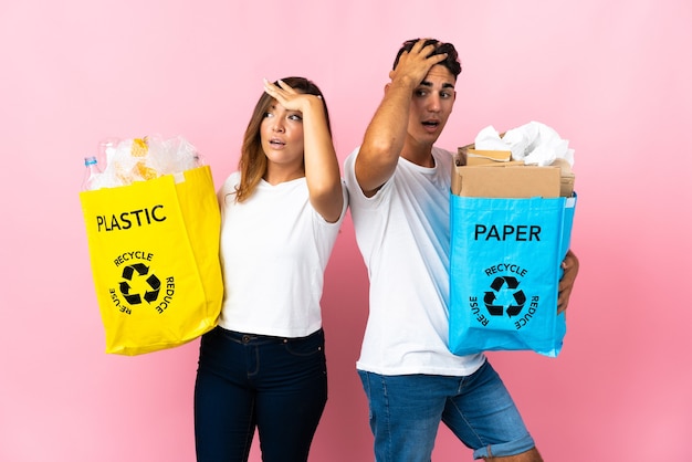 Young couple holding a bag full of plastic and paper on pink doing surprise gesture while looking to the side