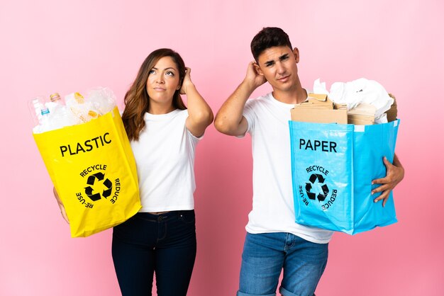 Young couple holding a bag full of plastic and paper on pink having doubts