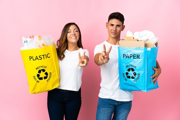 Foto giovani coppie che tengono un sacchetto pieno di plastica e carta sul rosa che sorride e che mostra il segno di vittoria