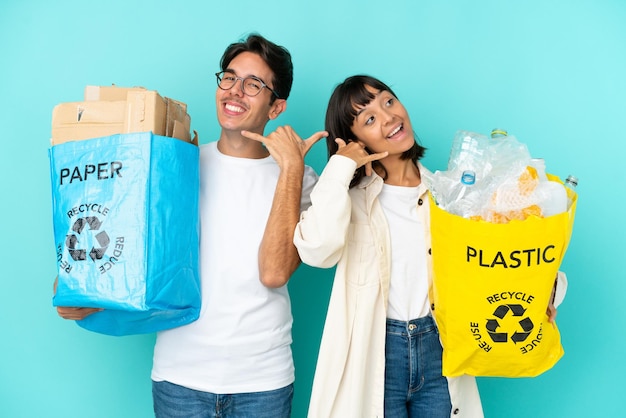 プラスチックと紙でいっぱいのバッグを持ってリサイクルする若いカップルは、青い背景で隔離され、電話ジェスチャーを作成します。