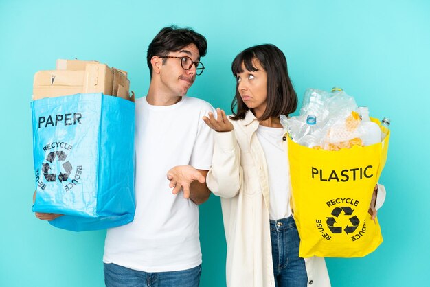 Young couple holding a bag full of plastic and paper to recycle isolated on blue background making unimportant gesture while lifting the shoulders