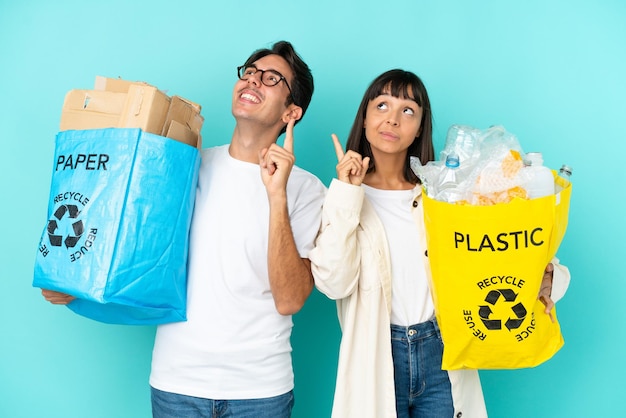 プラスチックと紙でいっぱいのバッグを持ってリサイクルする若いカップルは、人差し指で指している青い背景で隔離の素晴らしいアイデア