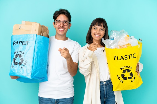 Young couple holding a bag full of plastic and paper to recycle isolated on blue background points finger at you with a confident expression