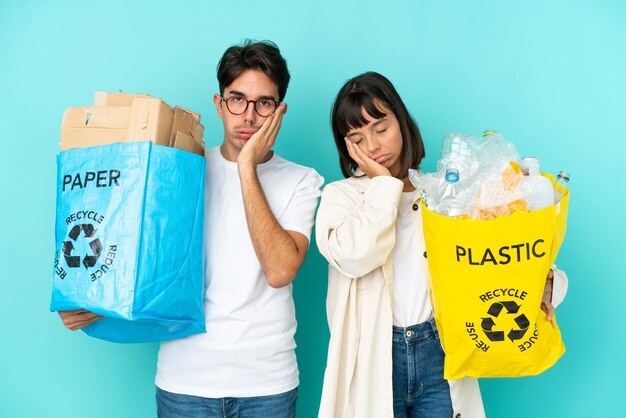 驚きとショックを受けた表情で青い背景に分離されたリサイクルするためにプラスチックと紙でいっぱいのバッグを保持している若いカップル