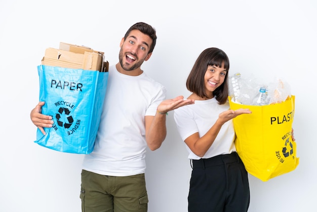 プラスチックと紙でいっぱいのバッグを持ってリサイクルする若いカップルは、白い背景で隔離され、手を横に伸ばして招待します