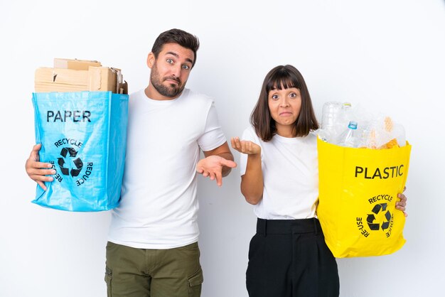 Young couple holding a bag full of plastic and paper to recycle isolated on white background having doubts while raising hands and shoulders