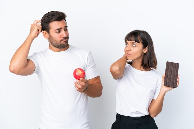 Young couple holding chocolate and apple isolated on white background having doubts while scratching head