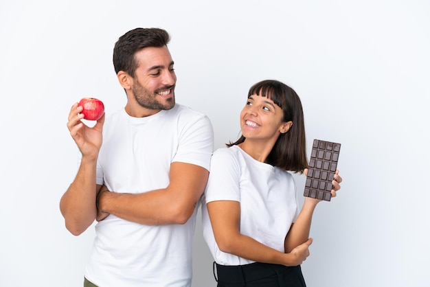 Young couple holding chocolate and apple isolated on white background looking over the shoulder with a smile