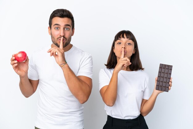 Young couple holding chocolate and apple isolated on white background showing a sign of silence gesture putting finger in mouth