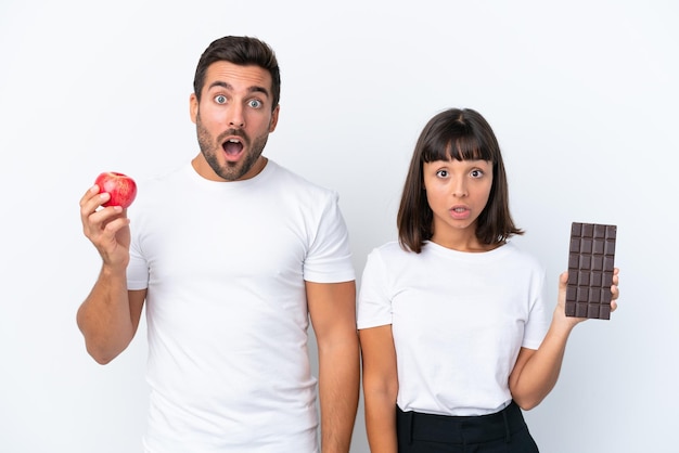 Young couple holding chocolate and apple isolated on white background with surprise and shocked facial expression