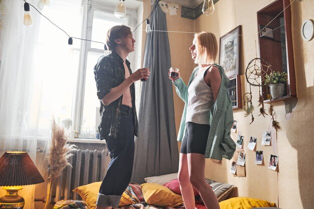 Photo young couple holding glasses of red wine standing on the bed and dancing together during domestic party