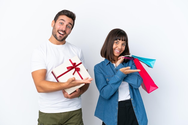 Young couple holding shopping bags and present isolated on white background extending hands to the side for inviting to come