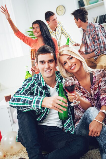 Young couple at home party toasting with drinks, smiling and looking at camera. In the background their friends talking.