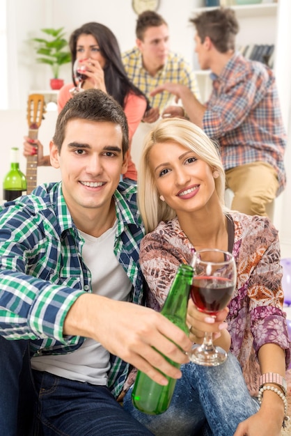 Young couple at home party with a smile on their faces,
toasting with drinks that are in the foreground. in the background
is they friends.