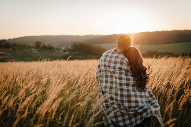 Young couple hugging, standing back, people covered with blanket, at sunset in autumn outdoor. at field grass on background of sun. full length. Close Up.