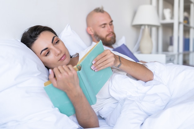 Young couple husband with wife at home in bed reading books in autumn winter evening, man fell asleep