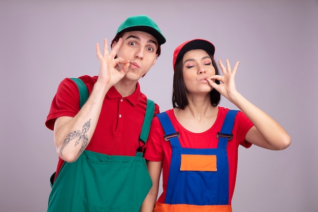 Young couple impressed guy pleased girl in construction worker uniform and cap guy  zipping his mouth with gesture girl doing delicious gesture with closed eyes