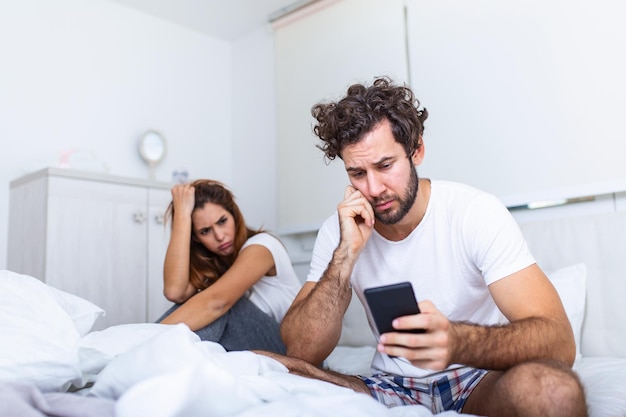 若いカップルがベッドに座っています。男は彼のスマートフォンで何かを見ています。その少女は彼に腹を立てている。彼女は電話で彼氏にイライラして欲求不満になっている彼を見ています
