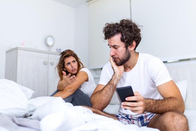 写真 若いカップルがベッドに座っています。男は彼のスマートフォンで何かを見ています。その少女は彼に腹を立てている。彼女は電話で彼氏にイライラして欲求不満になっている彼を見ています