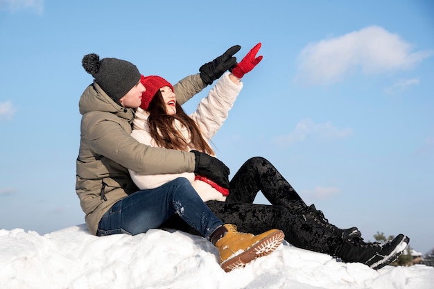 Young couple is sitting in snow and pointing to the sky.