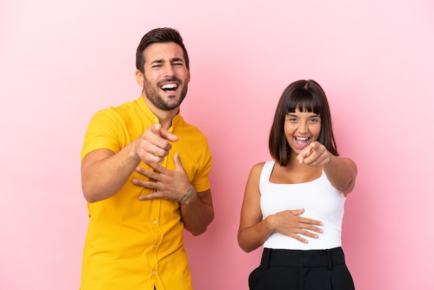 Young couple isolated on pink background pointing with finger at someone and laughing a lot