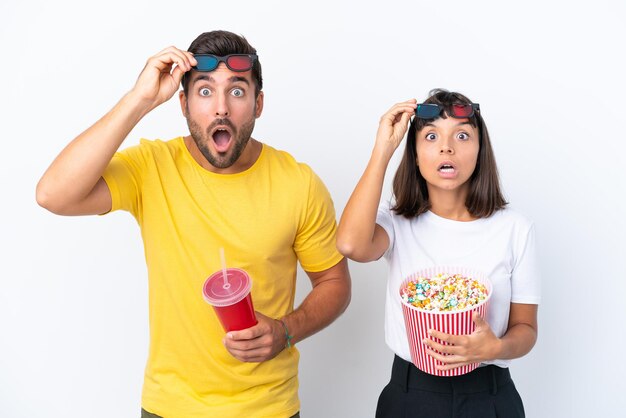 Young couple isolated on white background surprised with 3d glasses and holding a big bucket of popcorns