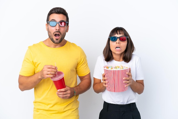 Young couple isolated on white background surprised with 3d glasses and holding a big bucket of popcorns